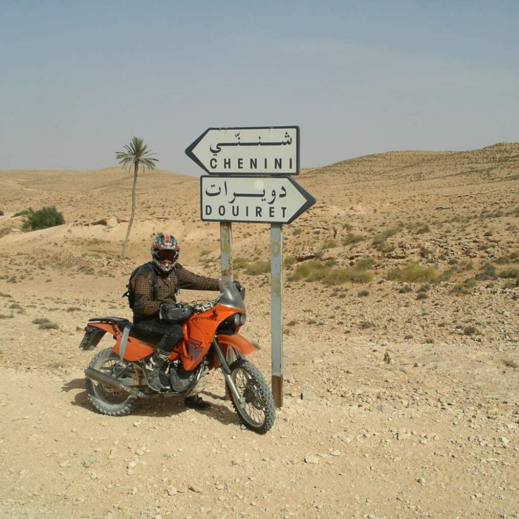 Un motociclista con una moto enduro arancione accanto a un cartello per Chenini e Douiret